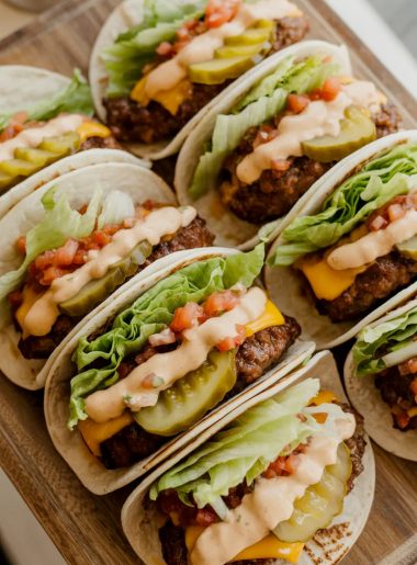Six smash burger tacos on a wooden serving board loaded with melted cheddar, shredded lettuce, diced tomatoes, pickles, and special sauce drizzle