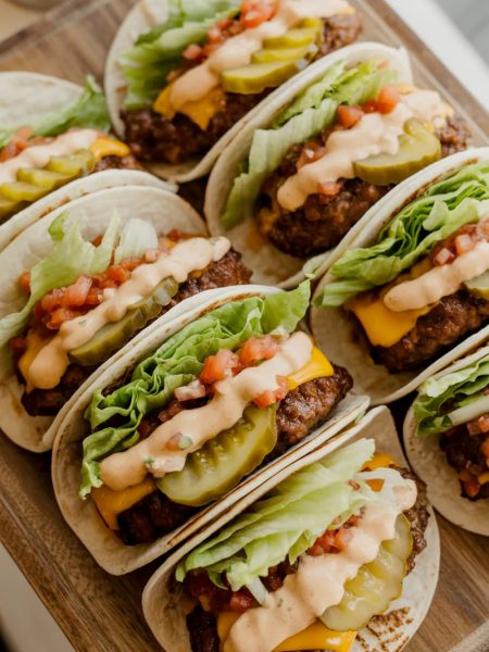 Six smash burger tacos on a wooden serving board loaded with melted cheddar, shredded lettuce, diced tomatoes, pickles, and special sauce drizzle
