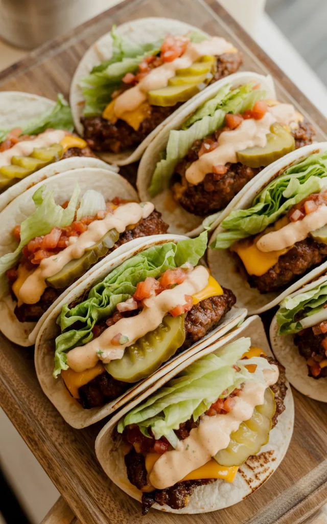 Six smash burger tacos on a wooden serving board loaded with melted cheddar, shredded lettuce, diced tomatoes, pickles, and special sauce drizzle
