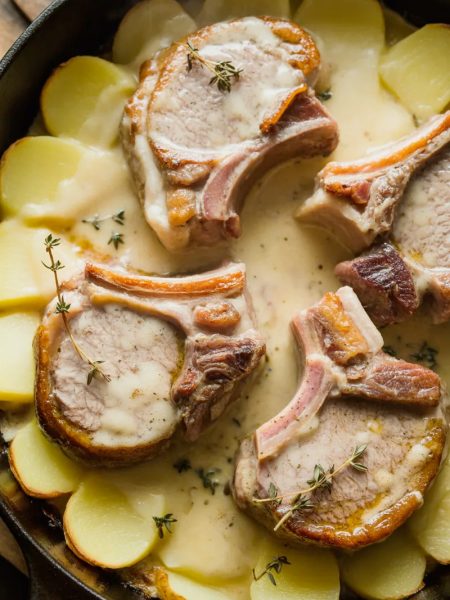 Overhead close-up of golden seared pork chops nestled into creamy scalloped potatoes in a cast iron skillet garnished with fresh thyme sprigs