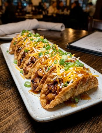A plate of crispy chicken wonton tacos drizzled with sweet chili sauce, topped with shredded cheese and sliced green onions, served on a white platter