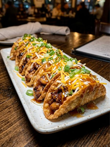 A plate of crispy chicken wonton tacos drizzled with sweet chili sauce, topped with shredded cheese and sliced green onions, served on a white platter