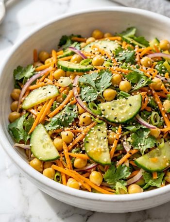 A close-up of a vibrant Asian cucumber and chickpea slaw with sliced cucumbers, shredded carrots, chickpeas, red onion, fresh cilantro, and sesame seeds tossed in a bold sesame ginger dressing served in a large white bowl on a marble surface