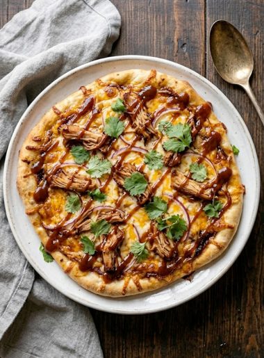 BBQ chicken flatbread pizza topped with shredded chicken melted mozzarella and cheddar cheese red onion slices drizzled BBQ sauce and fresh cilantro on a white plate