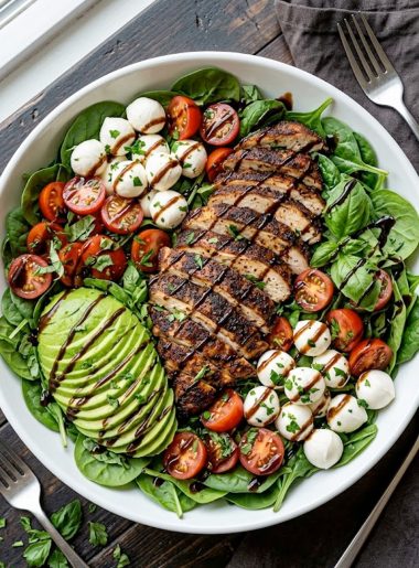 A large white ceramic bowl filled with fresh baby spinach topped with sliced smoky baked marinated chicken fresh mozzarella balls halved cherry tomatoes sliced avocado fresh basil and a generous drizzle of balsamic vinaigrette