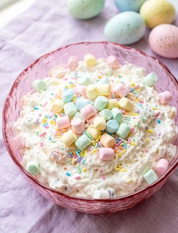 A top-down view of a creamy Easter fluff salad loaded with colorful pastel mini marshmallows and Easter sprinkles served in a pink glass bowl on a lavender linen with speckled Easter eggs in the background