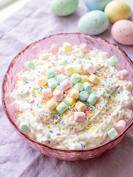 A top-down view of a creamy Easter fluff salad loaded with colorful pastel mini marshmallows and Easter sprinkles served in a pink glass bowl on a lavender linen with speckled Easter eggs in the background