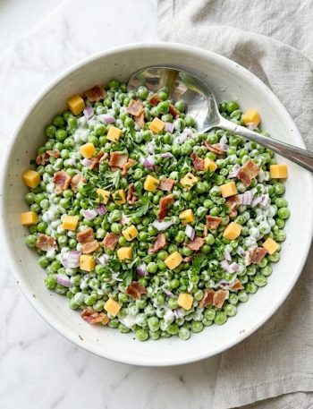A top-down view of a vibrant spring pea salad packed with sweet green peas, crumbled crispy bacon, diced sharp cheddar cheese, finely diced red onion, and fresh herbs all coated in a creamy tangy dressing in a large white bowl on a white marble surface