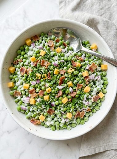 A top-down view of a vibrant spring pea salad packed with sweet green peas, crumbled crispy bacon, diced sharp cheddar cheese, finely diced red onion, and fresh herbs all coated in a creamy tangy dressing in a large white bowl on a white marble surface
