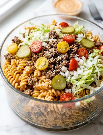 A large glass bowl of Big Mac pasta salad loaded with rotini pasta ground beef cherry tomatoes pickles lettuce and sesame seeds with creamy Big Mac sauce