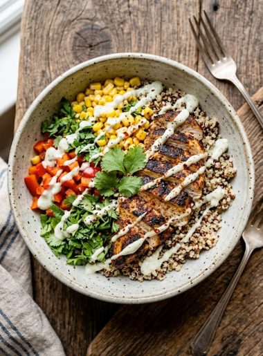 A bowl of blackened chicken quinoa with sweet corn, fresh herbs, and creamy sauce drizzled on top, served in a gray ceramic bowl