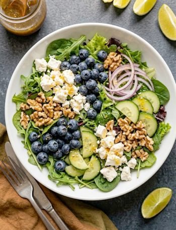 Overhead view of a blueberry Easter salad with crumbled feta, walnuts, cucumber, and mixed greens in a wide white bowl drizzled with honey lemon vinaigrette