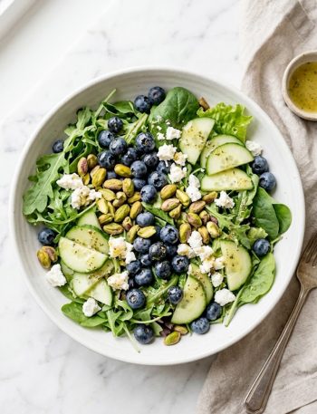 A vibrant blueberry pistachio spring salad with goat cheese, cucumber, and fresh greens in a white bowl