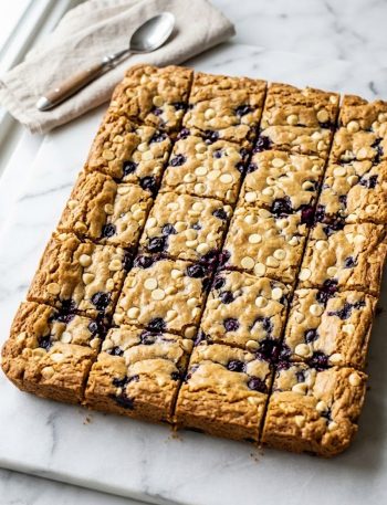 A close-up overhead view of golden blueberry white chocolate blondies cut into squares showing jammy blueberry pockets and white chocolate chips throughout the crackly golden surface