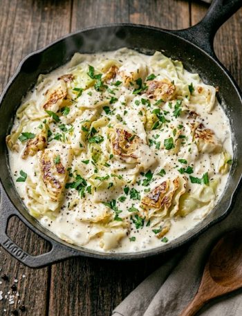 A close-up overhead shot of creamy cabbage Alfredo in a large black skillet with tender cabbage leaves coated in a rich white Alfredo sauce garnished with fresh parsley and cracked black pepper