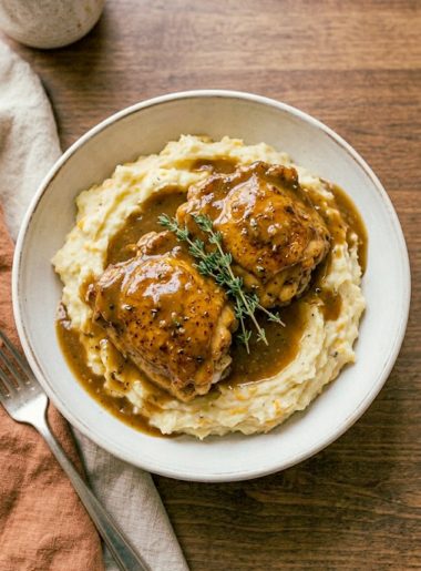 A wide ceramic bowl of golden seared chicken thighs smothered in rich garlic gravy served over a generous mound of creamy cheesy mashed potatoes garnished with fresh thyme