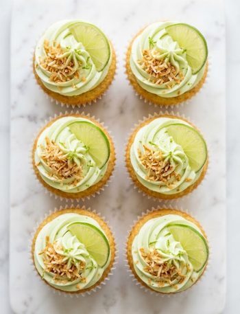 A plate of coconut cupcakes with lime frosting topped with toasted coconut flakes and fresh lime slices on a white marble surface