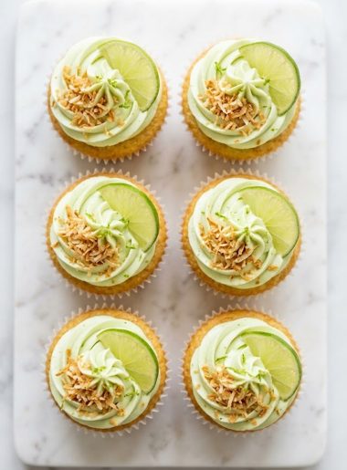A plate of coconut cupcakes with lime frosting topped with toasted coconut flakes and fresh lime slices on a white marble surface