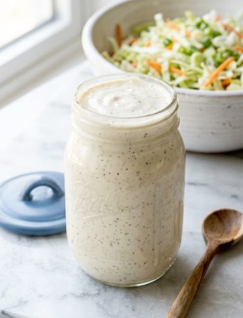 A glass mason jar filled with thick creamy white coleslaw dressing sitting in front of a large bowl of shredded cabbage and carrot coleslaw mix on a light marble surface with a wooden spoon beside it