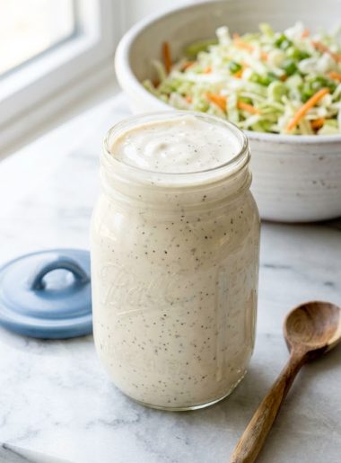 A glass mason jar filled with thick creamy white coleslaw dressing sitting in front of a large bowl of shredded cabbage and carrot coleslaw mix on a light marble surface with a wooden spoon beside it