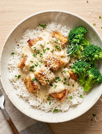 A bowl of creamy garlic chicken rice with steamed broccoli and fresh parsley garnish on a rustic wooden surface