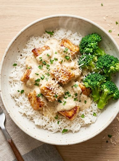 A bowl of creamy garlic chicken rice with steamed broccoli and fresh parsley garnish on a rustic wooden surface