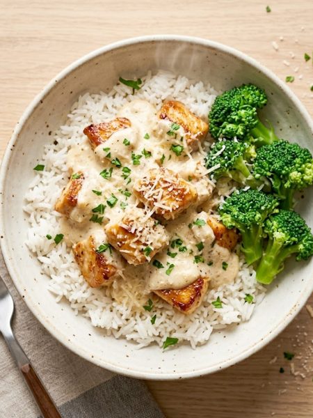 A bowl of creamy garlic chicken rice with steamed broccoli and fresh parsley garnish on a rustic wooden surface