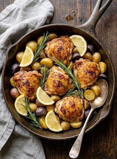 Crispy lemon herb chicken thighs with roasted baby potatoes in a cast iron skillet garnished with fresh rosemary and lemon slices