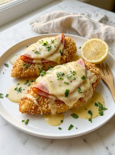 A close-up of golden crispy breaded Malibu chicken topped with savory ham, melted Swiss cheese, and a drizzle of creamy honey mustard sauce garnished with fresh parsley and a lemon wedge on a white plate
