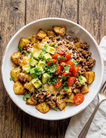 A white bowl filled with crispy roasted potato chunks topped with seasoned ground beef, melted cheese, diced avocado, cherry tomatoes, and fresh cilantro
