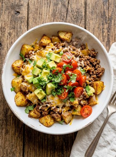 A white bowl filled with crispy roasted potato chunks topped with seasoned ground beef, melted cheese, diced avocado, cherry tomatoes, and fresh cilantro