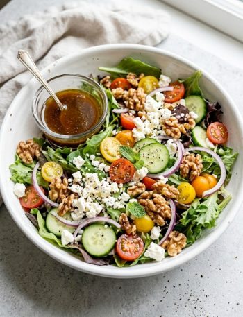 Easy spring mix salad with cherry tomatoes cucumber red onion crumbled feta and toasted walnuts in a white bowl with a small glass of balsamic honey dressing on the side