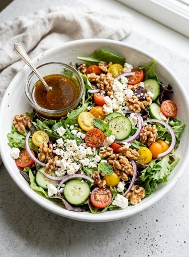 Easy spring mix salad with cherry tomatoes cucumber red onion crumbled feta and toasted walnuts in a white bowl with a small glass of balsamic honey dressing on the side