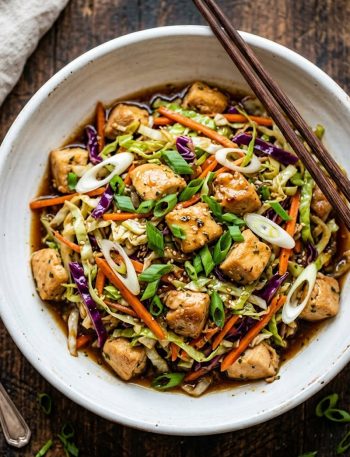 A white ceramic bowl filled with savory egg roll bowl featuring diced chicken shredded green and purple cabbage carrots and green onions in a glossy soy ginger garlic sauce on a rustic wooden surface