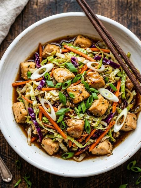 A white ceramic bowl filled with savory egg roll bowl featuring diced chicken shredded green and purple cabbage carrots and green onions in a glossy soy ginger garlic sauce on a rustic wooden surface