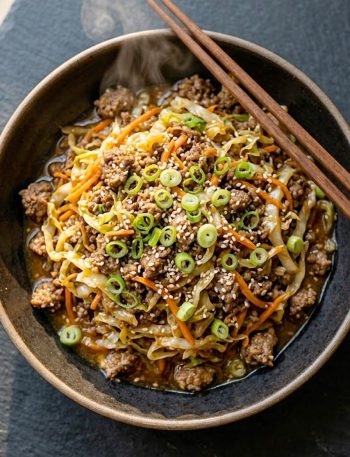A bowl of egg roll in a bowl with seasoned ground beef, wilted cabbage, carrots, green onions, and sesame seeds on a dark slate surface.