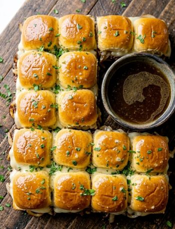 Golden French dip sliders on a wooden board with a small bowl of beef broth dipping sauce on the side