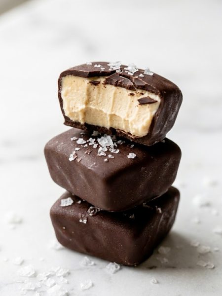 A stack of frozen peanut butter Greek yogurt bites coated in dark chocolate with flaky sea salt on top one bitten open to reveal the creamy peanut butter center