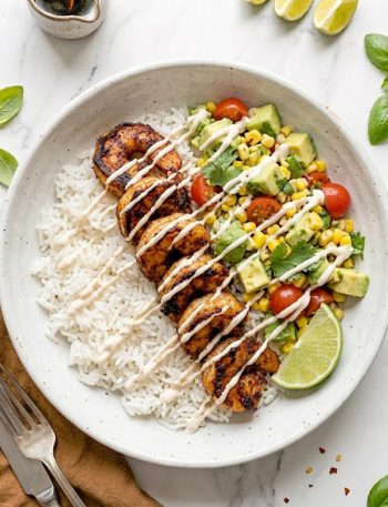 A white ceramic bowl filled with fluffy white rice topped with smoky seared garlic shrimp avocado corn salsa cherry tomatoes fresh cilantro and a creamy spicy mayo drizzle with a lime wedge on the side