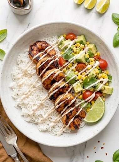 A white ceramic bowl filled with fluffy white rice topped with smoky seared garlic shrimp avocado corn salsa cherry tomatoes fresh cilantro and a creamy spicy mayo drizzle with a lime wedge on the side
