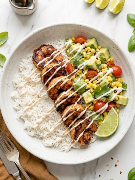 A white ceramic bowl filled with fluffy white rice topped with smoky seared garlic shrimp avocado corn salsa cherry tomatoes fresh cilantro and a creamy spicy mayo drizzle with a lime wedge on the side
