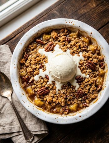 Overhead close-up of golden banana crumble topped with a melting scoop of vanilla ice cream in a white ceramic baking dish