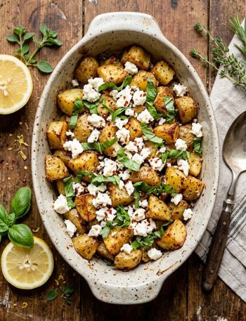 Crispy golden roasted potato chunks topped with crumbled white feta cheese and fresh basil leaves in a rustic baking dish
