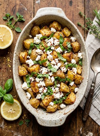 Crispy golden roasted potato chunks topped with crumbled white feta cheese and fresh basil leaves in a rustic baking dish