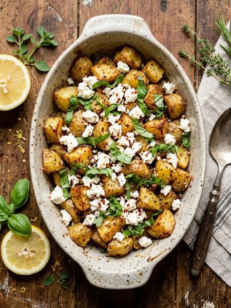 Crispy golden roasted potato chunks topped with crumbled white feta cheese and fresh basil leaves in a rustic baking dish