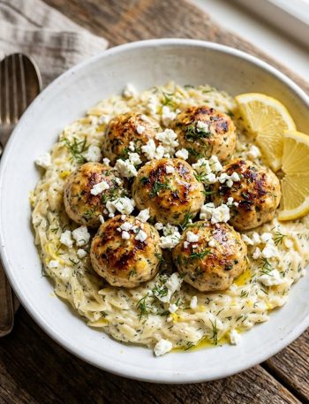 Golden seared Greek chicken meatballs nestled over creamy lemon orzo with crumbled feta and fresh dill in a white ceramic bowl on a rustic wooden surface