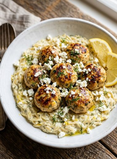 Golden seared Greek chicken meatballs nestled over creamy lemon orzo with crumbled feta and fresh dill in a white ceramic bowl on a rustic wooden surface