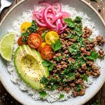 Overhead view of a ground beef chimichurri bowl with fluffy white rice, seasoned ground beef, sliced avocado, cherry tomatoes, pickled red onions, and a drizzle of bright green chimichurri sauce, garnished with fresh parsley and a lime wedge