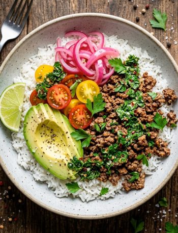 Overhead view of a ground beef chimichurri bowl with fluffy white rice, seasoned ground beef, sliced avocado, cherry tomatoes, pickled red onions, and a drizzle of bright green chimichurri sauce, garnished with fresh parsley and a lime wedge