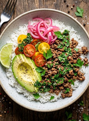 Overhead view of a ground beef chimichurri bowl with fluffy white rice, seasoned ground beef, sliced avocado, cherry tomatoes, pickled red onions, and a drizzle of bright green chimichurri sauce, garnished with fresh parsley and a lime wedge